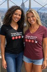 Two women standing side-by-side in front of a city skyline, wearing matching V-neck t-shirts with 'Times Square 42 St Station' and station number graphics printed on the front.