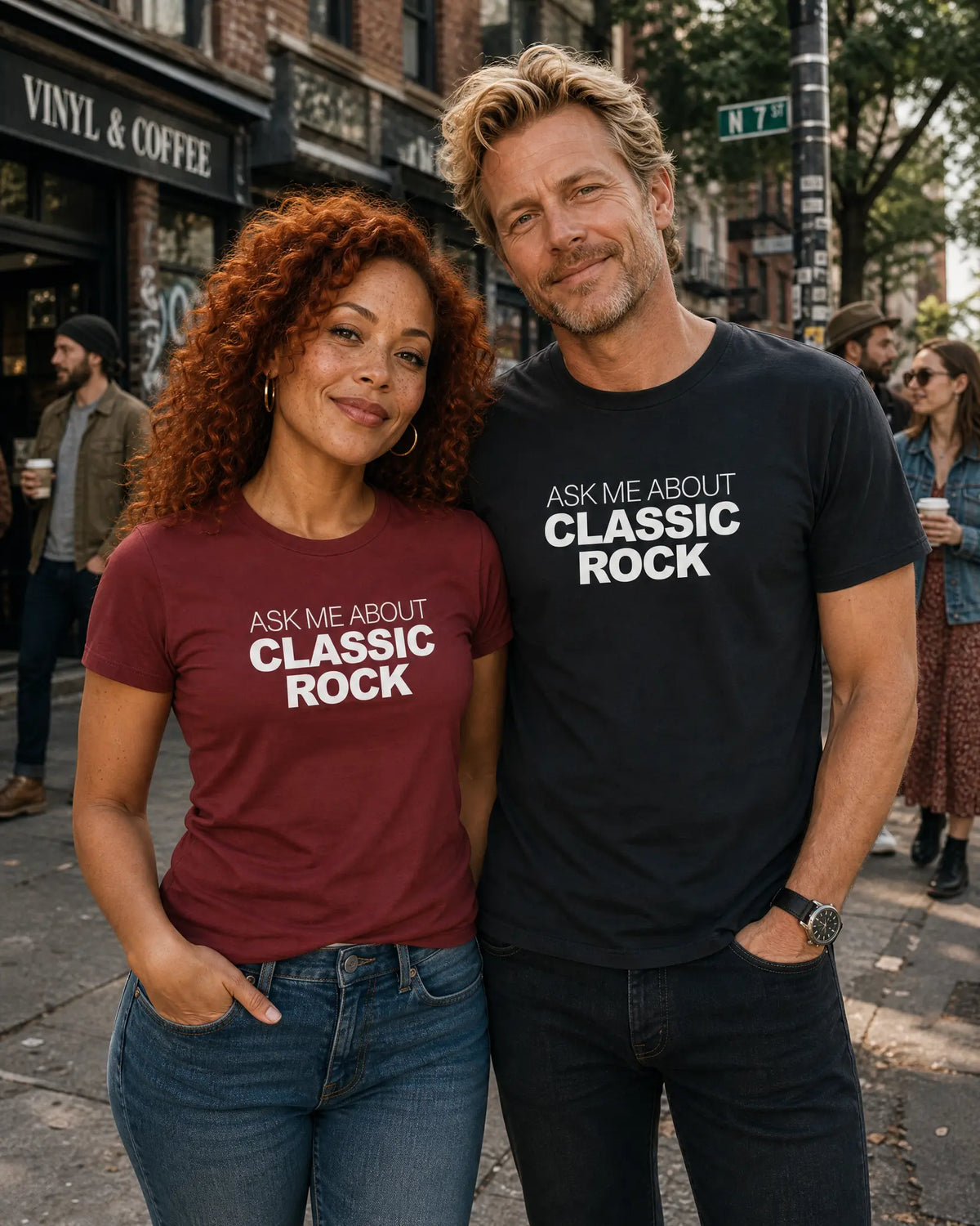 A man and a woman standing on a city street wearing matching graphic t-shirts with the text 'ASK ME ABOUT CLASSIC ROCK'. The woman's shirt is maroon, and the man's shirt is black.