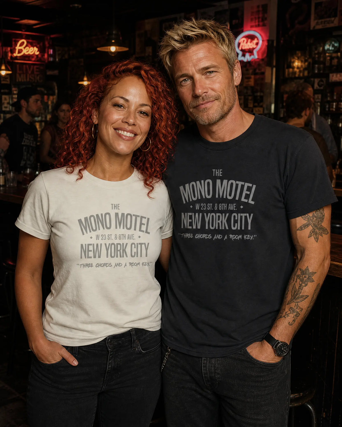 A smiling couple standing together in a dimly lit bar, both wearing matching graphic t-shirts featuring the text 'THE MONO MOTEL' and 'NEW YORK CITY'. The woman's shirt is a light heather gray, while the man's is black.