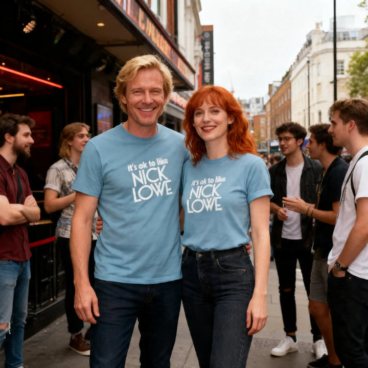 Two people wearing blue t-shirts with 'it's ok to like Nick Lowe' text on a street.